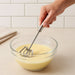 A person's hand holding a silver stainless steel hand whisk, mixing a pale yellow liquid in a clear glass bowl.
