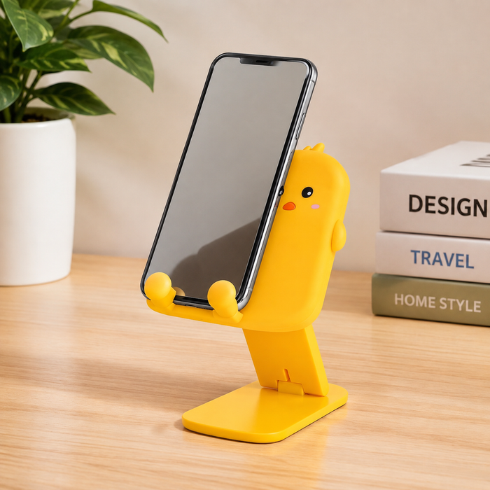 A yellow cartoon duck phone stand holds a smartphone on a wooden desk next to a plant and books.