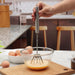 A person's hand holding a silver stainless steel hand whisk, mixing yellow liquid in a clear glass bowl on a wooden board.