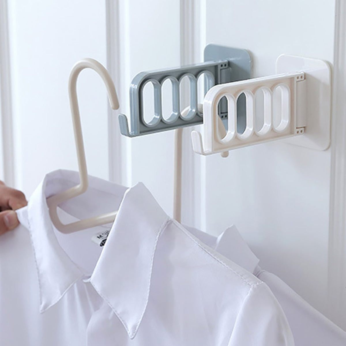 A hand places a white shirt on a hanger onto a white foldable wall hook, with a grey one next to it.