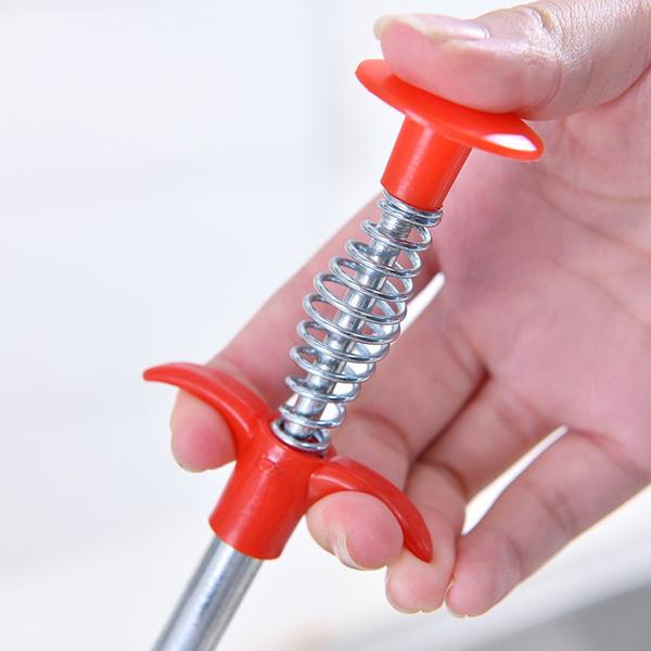 A hand pressing the red spring-loaded handle of the drain cleaning tool, showing the silver spring mechanism.