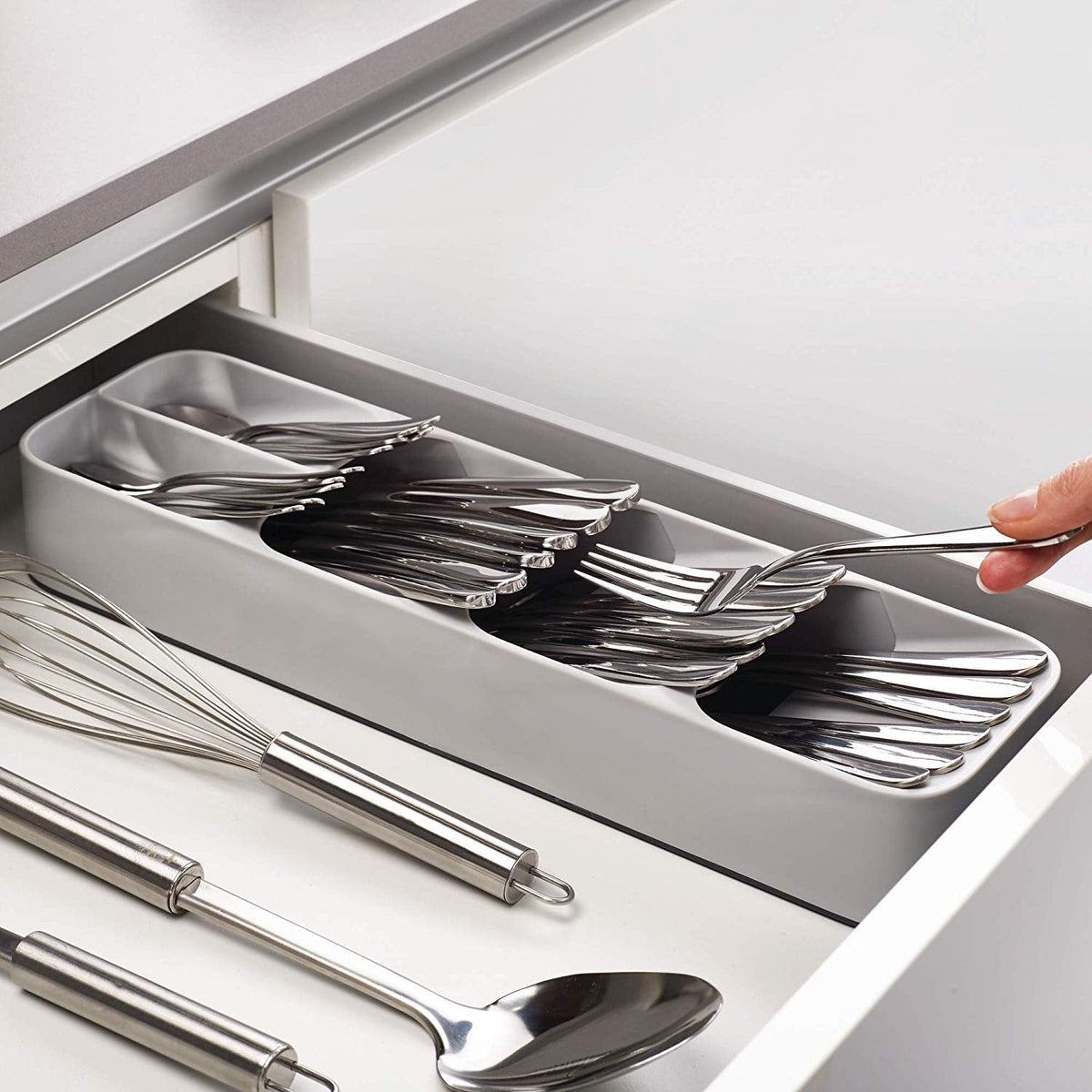 A hand places a fork into a gray plastic cutlery organizer filled with silverware inside a white kitchen drawer.