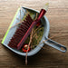 A red and white handled brush with brown bristles rests inside a gray dustpan filled with pine needles and small red