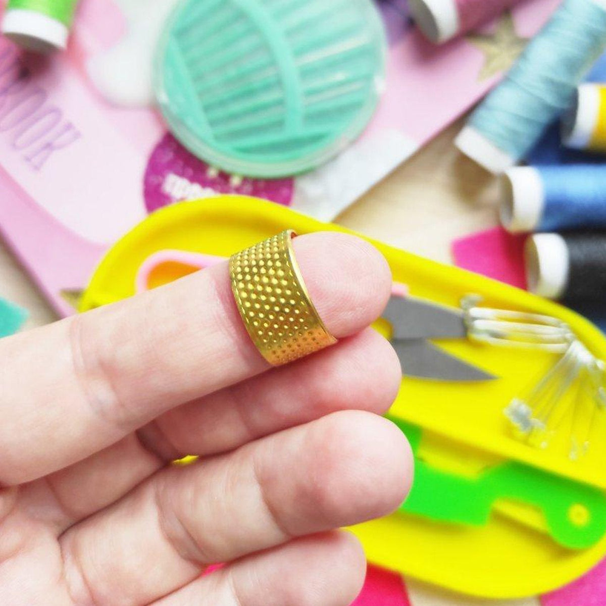 A person's finger wearing a gold-colored thimble with a textured surface, surrounded by blurred sewing items.
