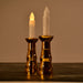 Two gold-based LED candles with illuminated flames stand on a dark surface in a dimly lit room.