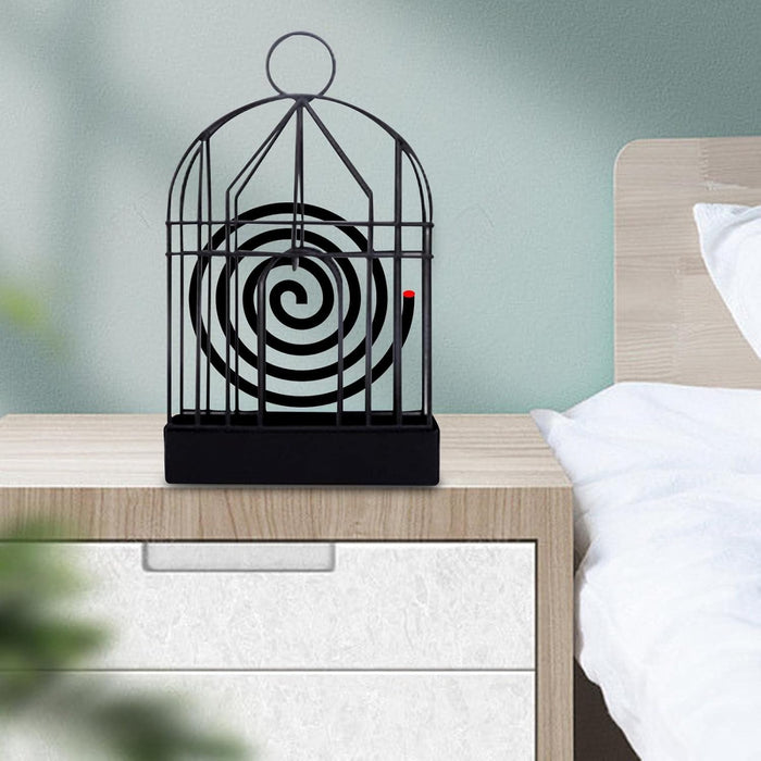A black iron birdcage-style holder with a burning mosquito coil sits on a light wood nightstand next to a bed.
