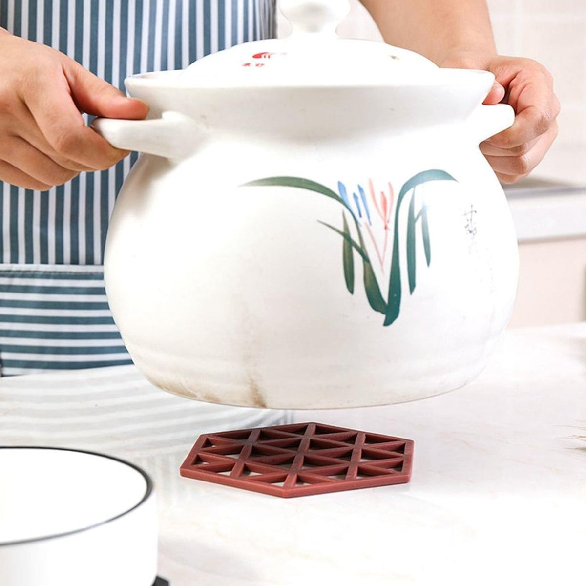 Hands hold a large white ceramic pot with green and red floral designs over a single maroon silicone coaster.