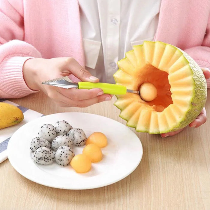 A person uses a green-handled tool to scoop a melon ball from a carved cantaloupe, with dragon fruit and melon balls on a