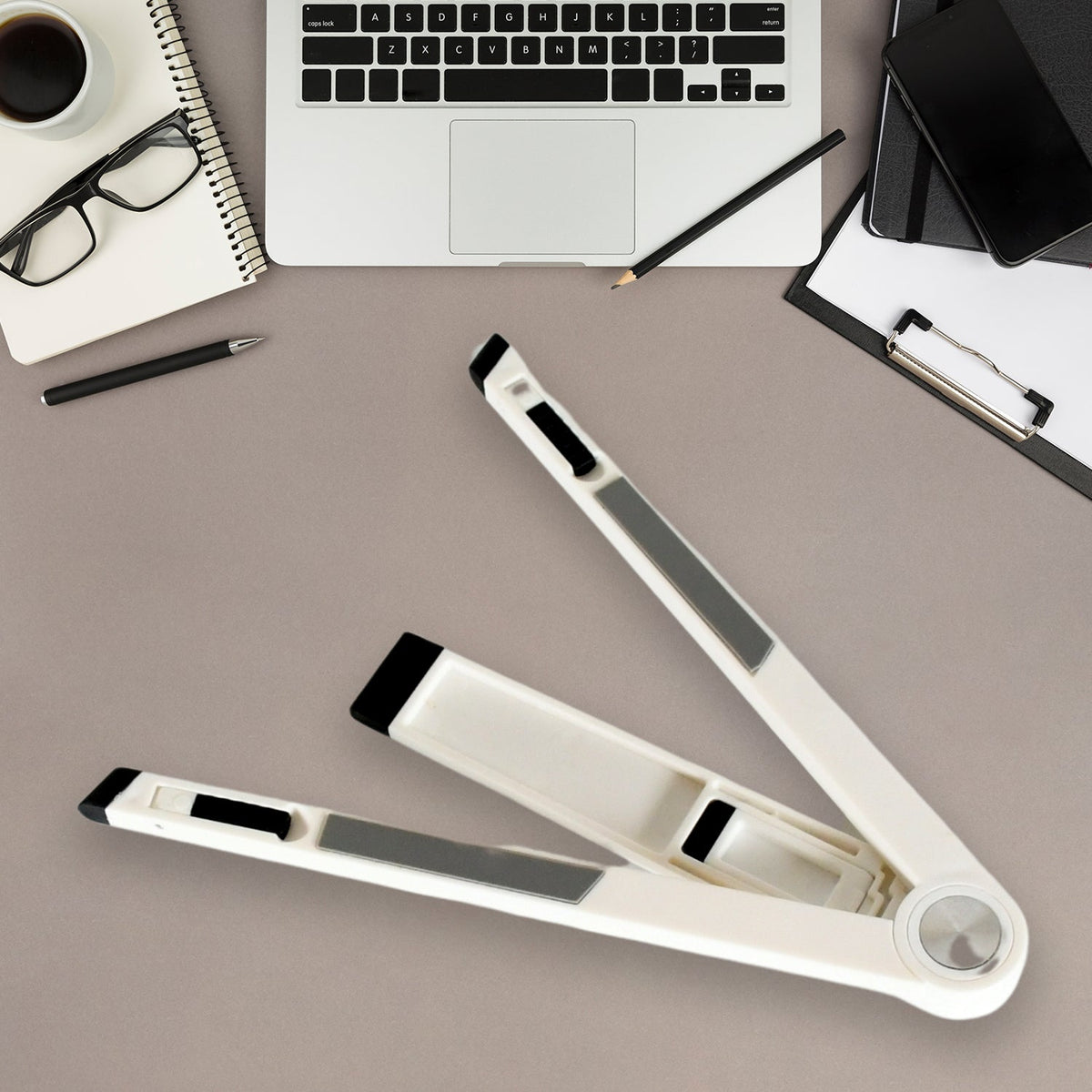 A white foldable stand, partially folded, rests on a gray desk with a laptop, notebook, and coffee.