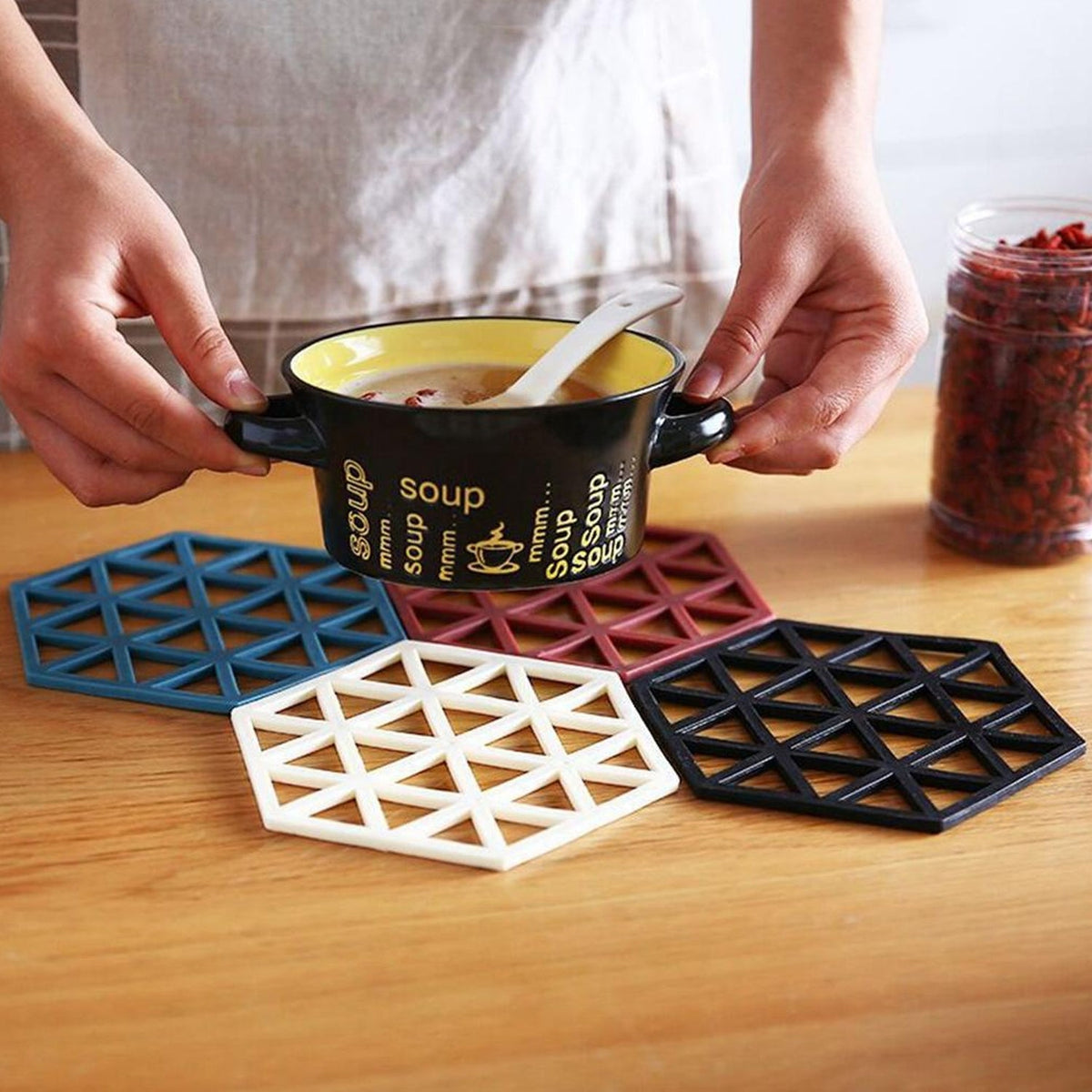 Hands hold a black bowl of soup over four hexagonal silicone coasters in blue, white, maroon, and black.