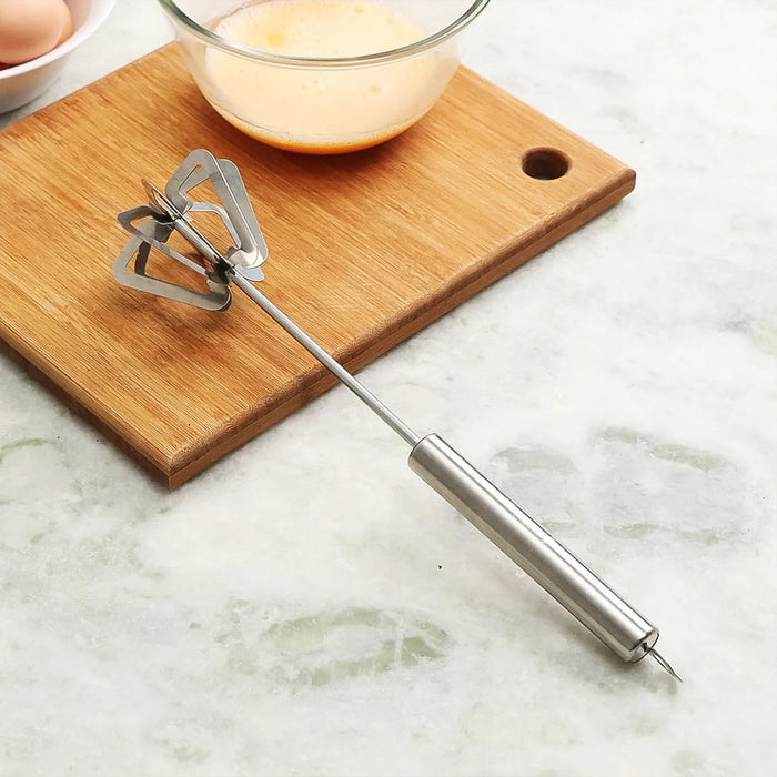 A silver stainless steel hand whisk resting on a brown wooden cutting board next to a glass bowl with yellow liquid.