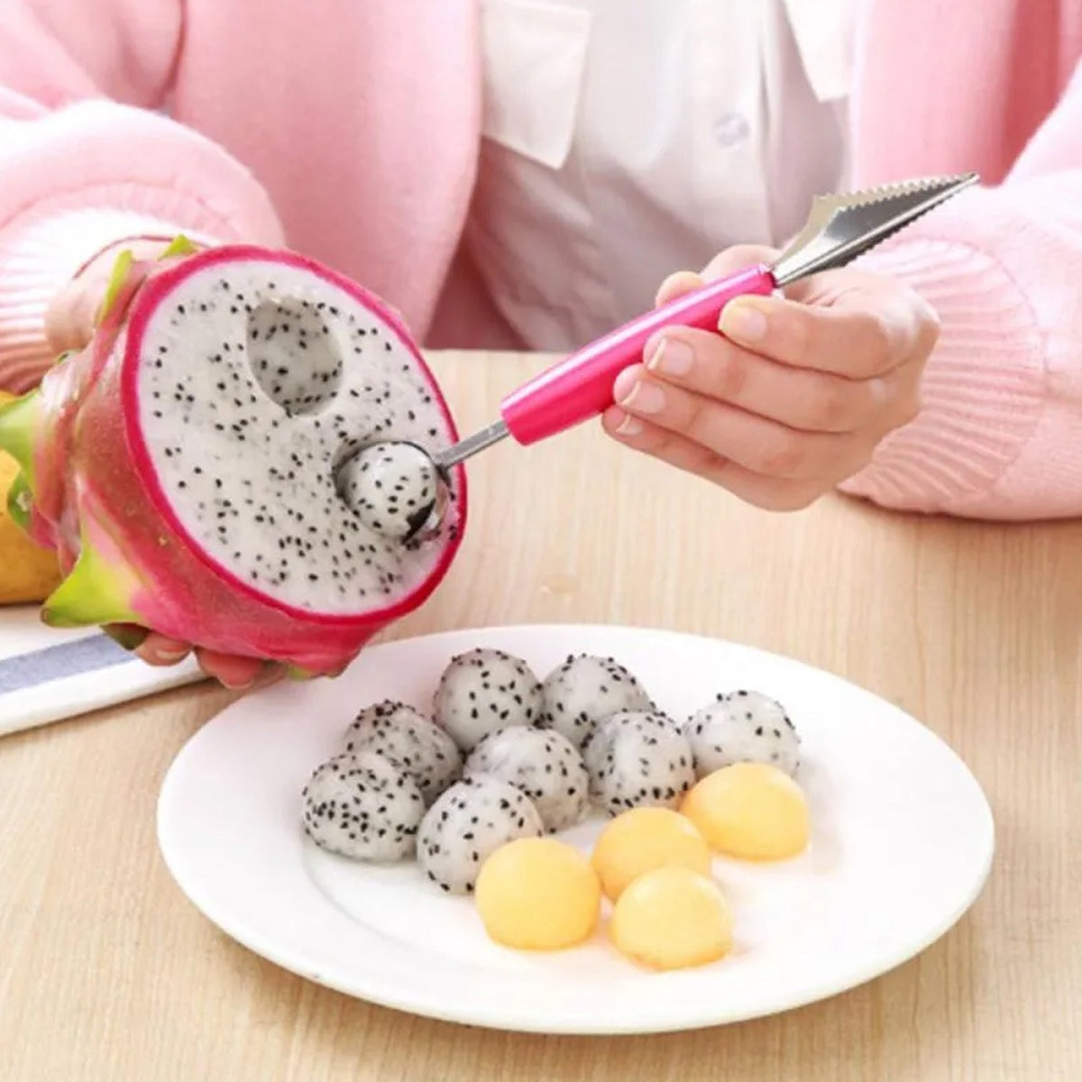 A person uses a pink-handled tool to scoop fruit from a halved dragon fruit, with dragon fruit and melon balls on a white