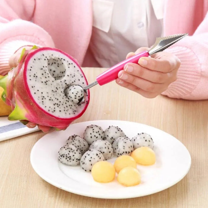 A person uses a pink-handled tool to scoop fruit from a halved dragon fruit, with dragon fruit and melon balls on a white