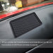 A black rectangular anti-skid mat is positioned on a car dashboard, with a wet car window in the background.