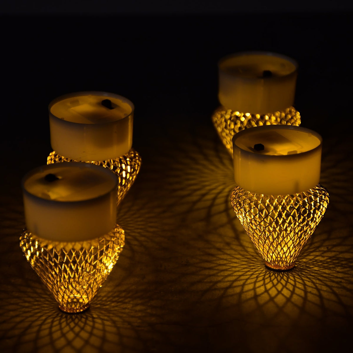 Four golden mesh LED tea lights casting intricate shadow patterns on a dark surface.