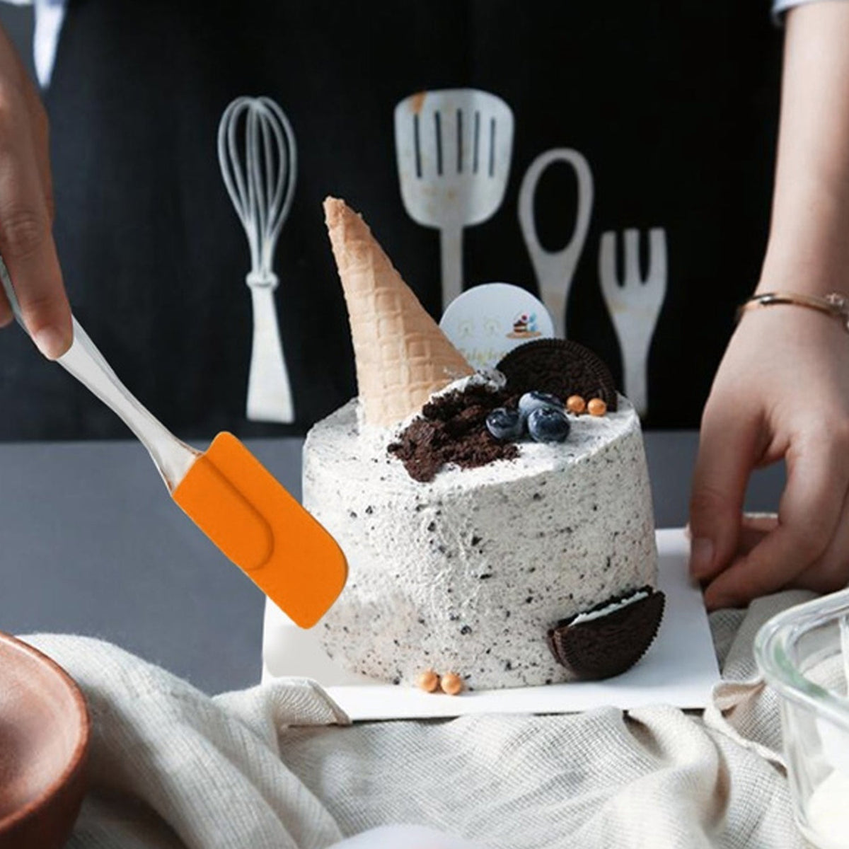 A hand uses an orange silicone spatula to serve a slice of a decorative white cake topped with an ice cream cone.
