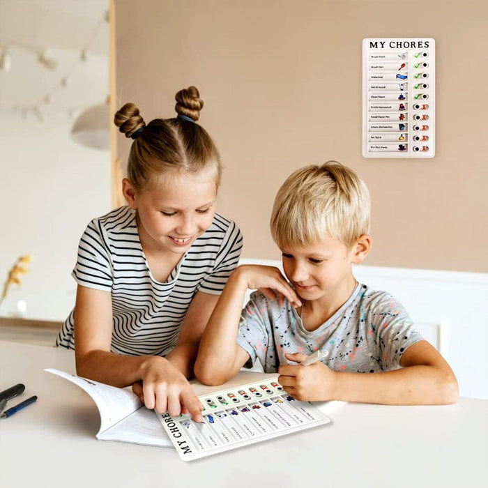 Two children sitting at a table, looking at a 'MY CHORES' checklist, with a board on the wall in the background.