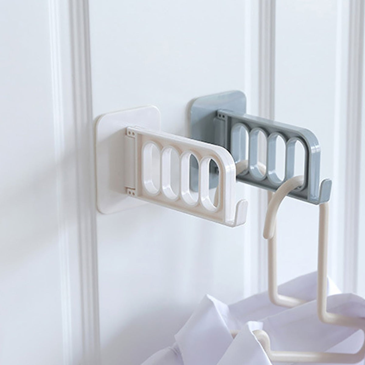 Two foldable wall hooks, one white and one grey, are mounted on a white door, holding shirts on hangers.
