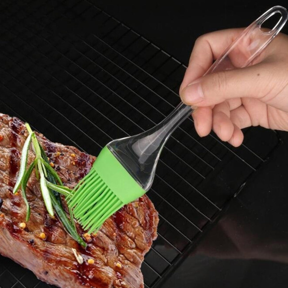 A hand uses a green silicone basting brush to spread sauce on a piece of grilled meat with rosemary sprigs.