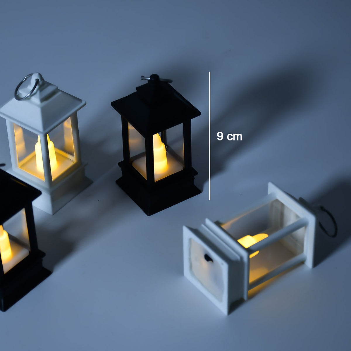 Four small black and white lanterns with glowing LED candles, one with a measurement of 9 cm indicated by a white line.