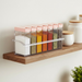 Six clear plastic spice jars with pink flip-top lids in a clear holder, filled with various spices, sitting on a wooden