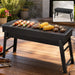 Black portable charcoal grill with food cooking on the grate, set outdoors on a wooden table.