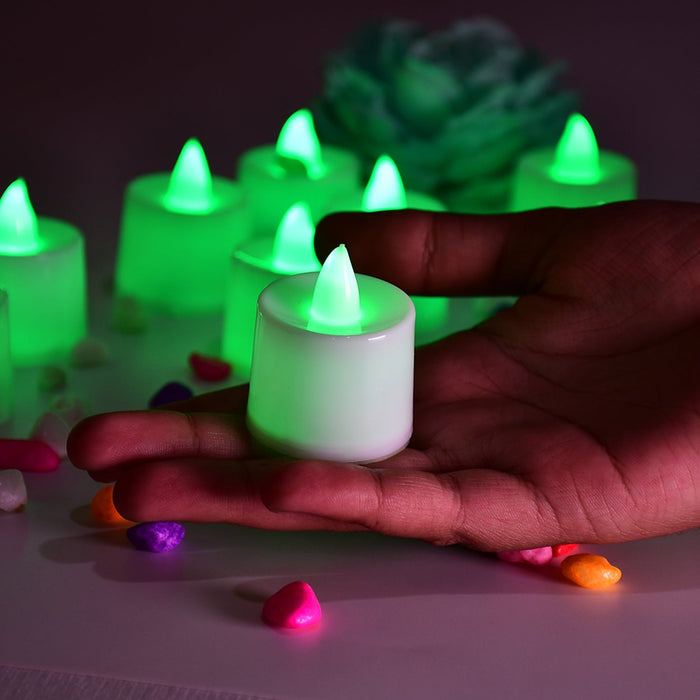 A hand gently holds a single green flameless LED tealight, surrounded by other glowing tealights and colorful pebbles.