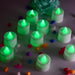 Ten green flameless LED tealights glow brightly on a white surface, surrounded by colorful decorative pebbles and a teal