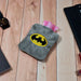 Gray plush hot water bag with a Batman logo on a wooden table with a watch and notebook.