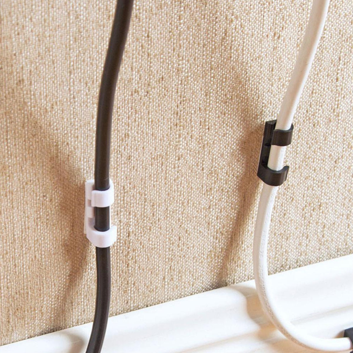Two cables, one black and one white, are secured to a textured beige wall with white and black self-adhesive clips.