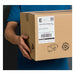 A person in a blue shirt holds a brown cardboard box with a large white shipping label affixed to its side.