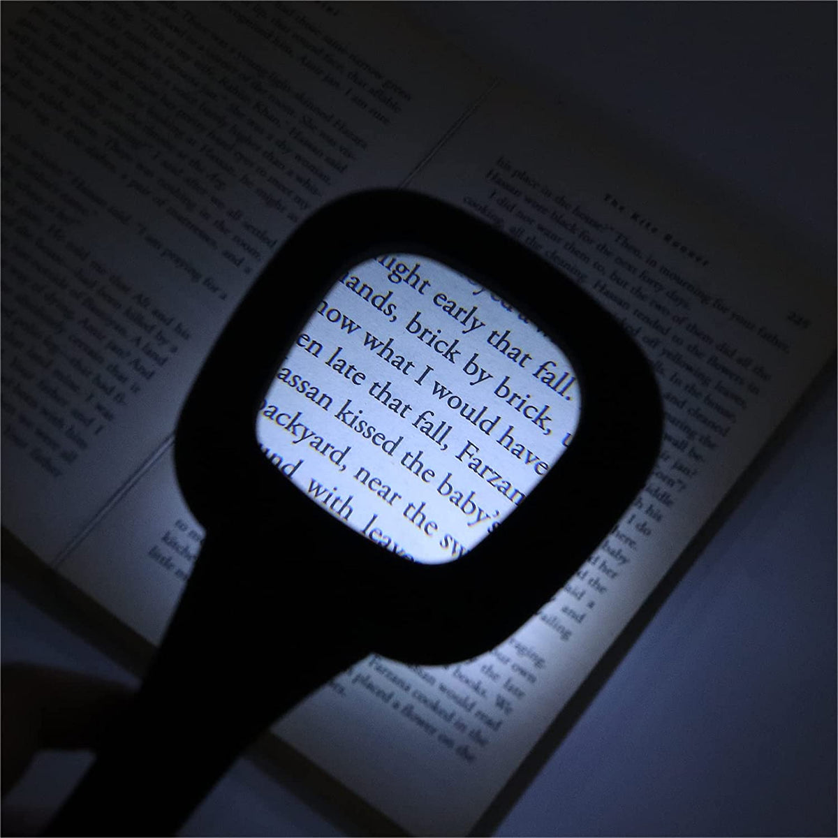 Black handheld magnifying glass with six LED lights illuminating text on an open book in a dimly lit setting.