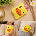 A yellow duck-shaped hot water bottle with a pink plastic insert on a marble surface with a candle and plant.