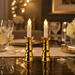 Two gold-based LED candles illuminate a dining table set with wine glasses and white flowers.