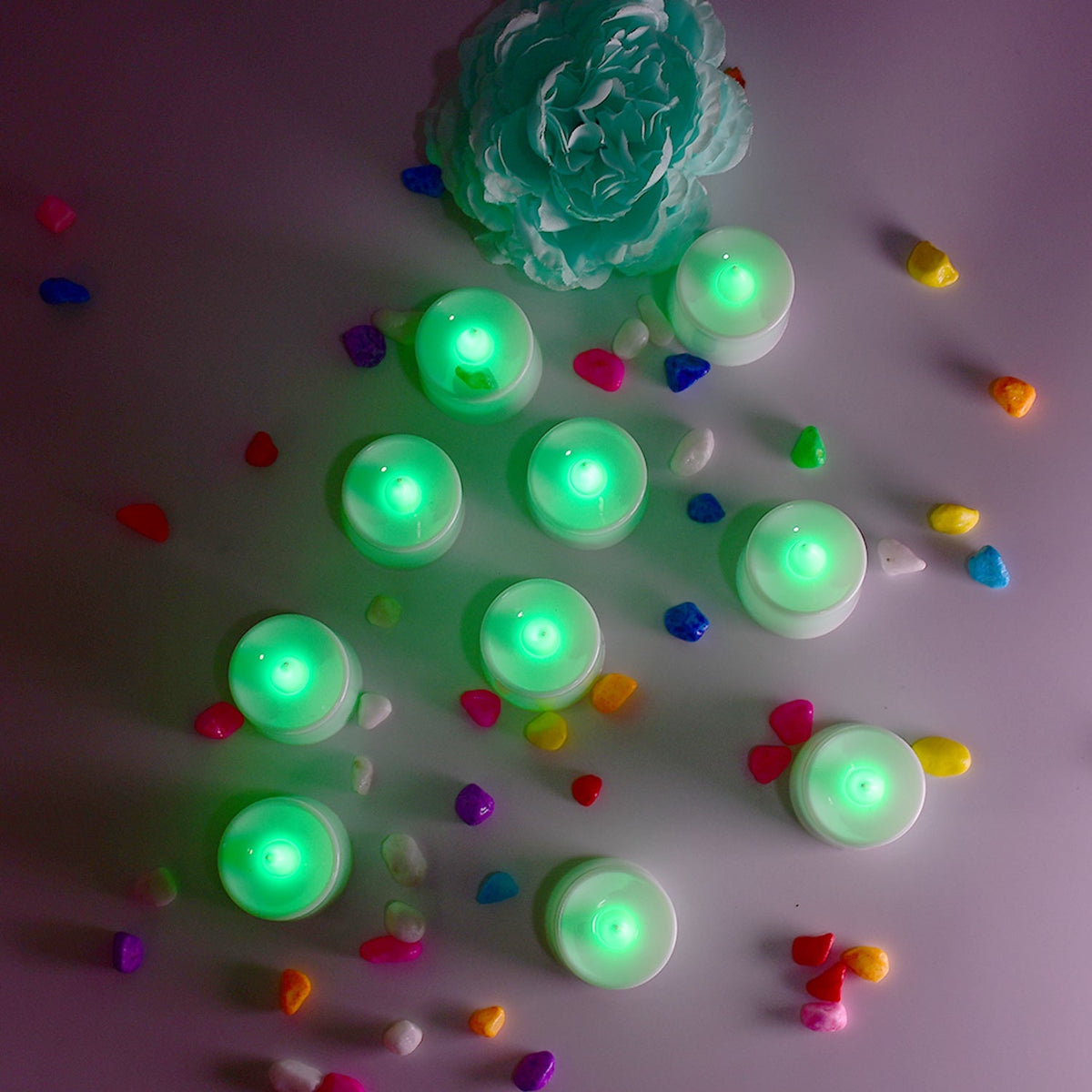 Ten green flameless LED tealights are scattered on a white surface with colorful pebbles and a teal decorative flower.