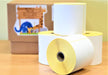 Four rolls of white thermal labels with yellow backing are stacked on a wooden surface, with a blurred brown box behind.