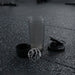 A disassembled grey and black shaker bottle with a metal whisk is on a dark gym floor.