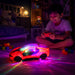 A young boy smiles while playing with a red remote control car that projects colorful lights onto the carpet in a dimly lit