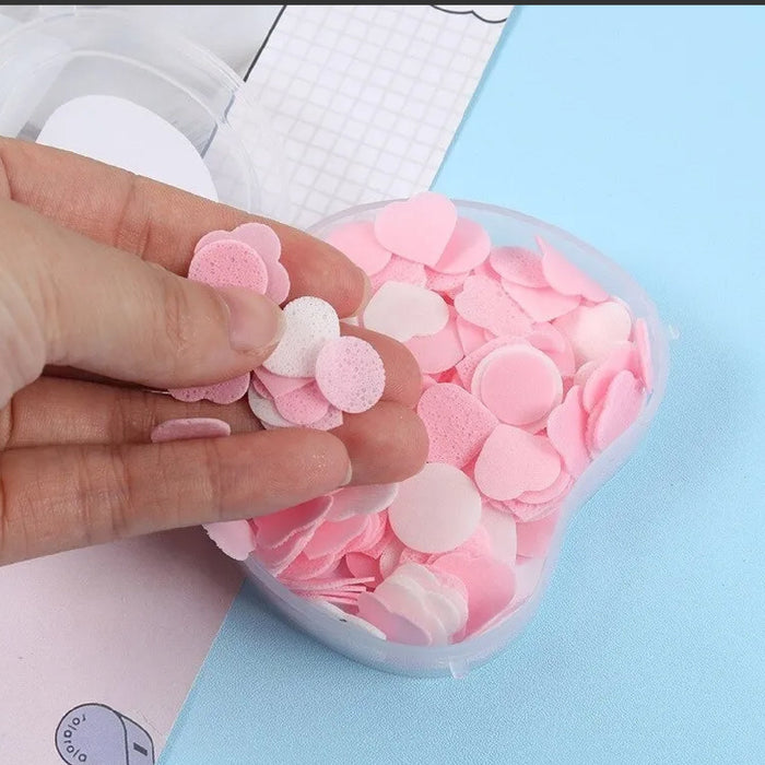A hand holds several pink and white heart and circle-shaped paper soap pieces above an open apple-shaped container.