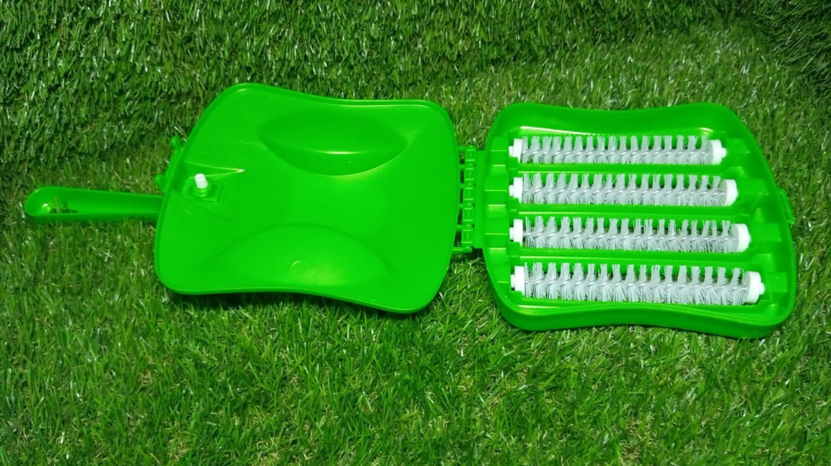 A bright green handheld carpet sweeper, open to reveal four rows of white brushes, resting on green artificial grass.