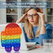 A rainbow Among Us character pop-it fidget toy is in the foreground, with a woman holding her temples in the background.